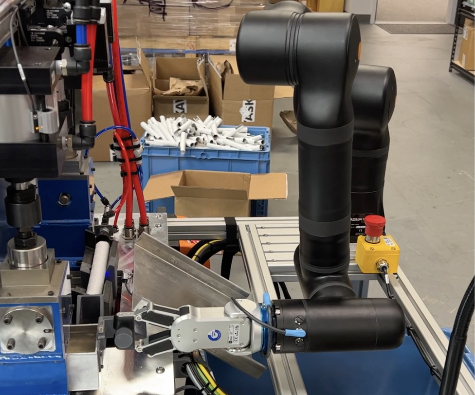 Industrial robotic arm and manufacturing equipment in warehouse workspace with blue storage bins and cardboard boxes