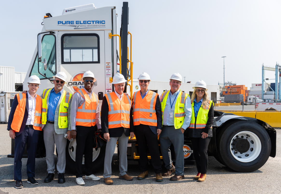 Team of eight workers in safety vests and hard hats posing before a Pure Electric truck at an industrial port facility