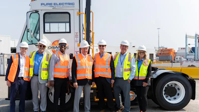 Team of eight workers in safety vests and hard hats posing before a Pure Electric truck at an industrial port facility