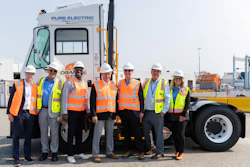Team of eight workers in safety vests and hard hats posing before a Pure Electric truck at an industrial port facility