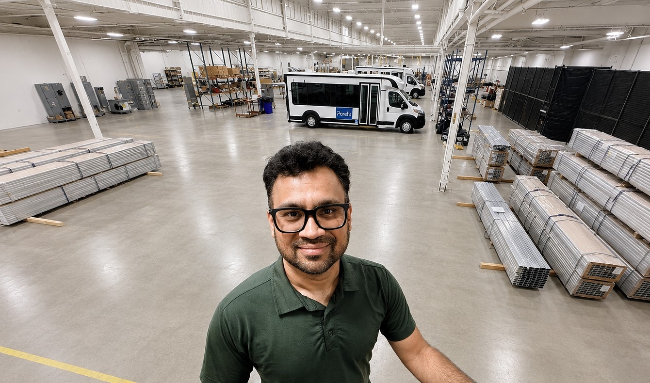 Man in glasses and green polo shirt smiling at camera in industrial warehouse with pallets and delivery van