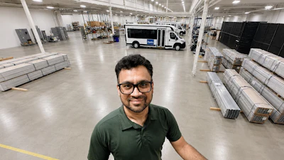 Man in glasses and green polo shirt smiling at camera in industrial warehouse with pallets and delivery van