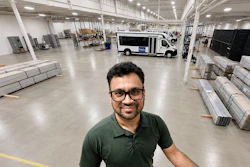 Man in glasses and green polo shirt smiling at camera in industrial warehouse with pallets and delivery van