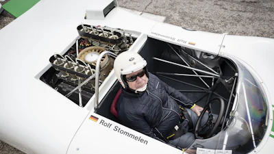 White race car with open cockpit and driver in helmet, viewed from above showing engine compartment