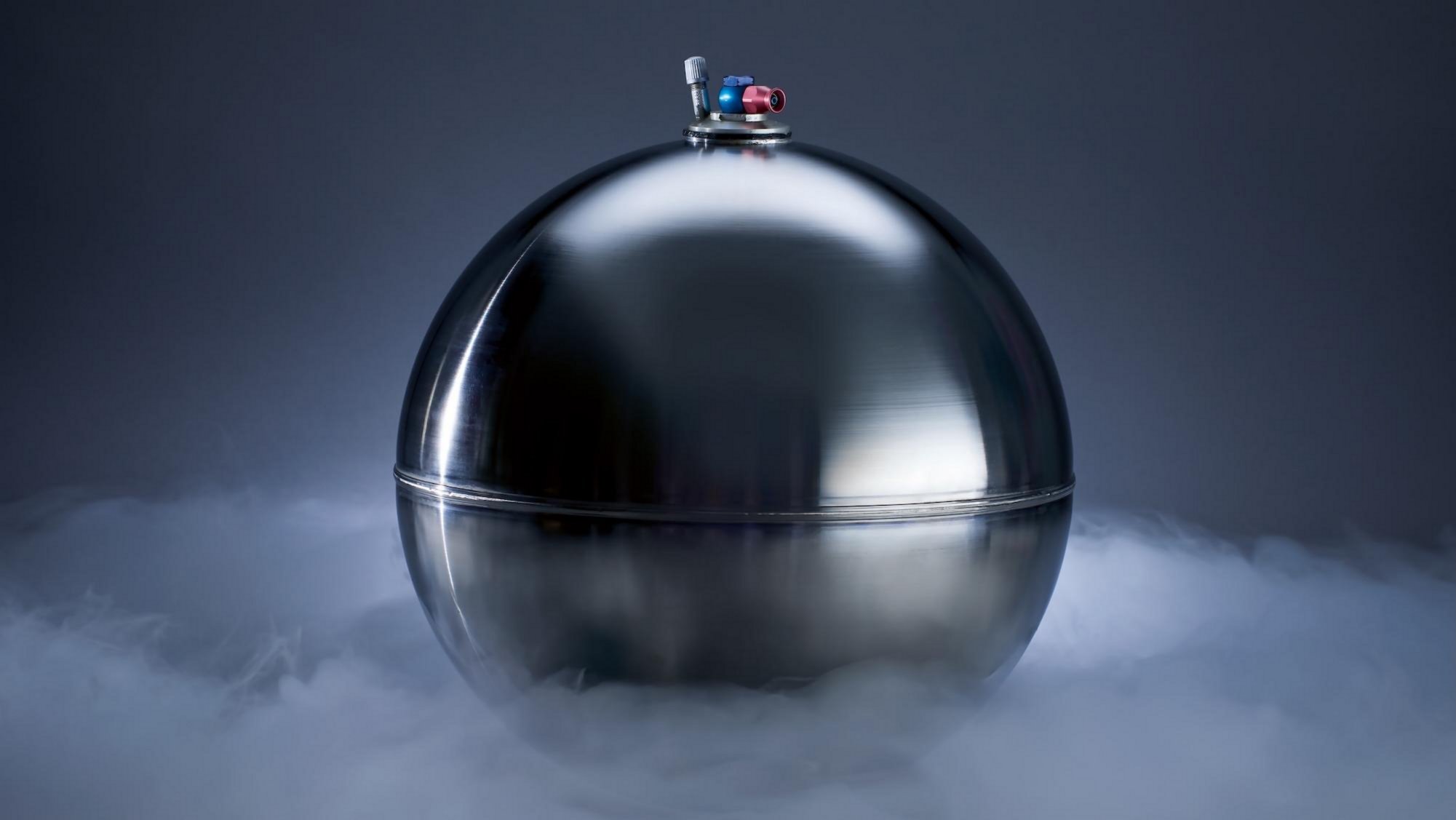 Polished silver spherical object with ornamental finial surrounded by dry ice vapor on dark background