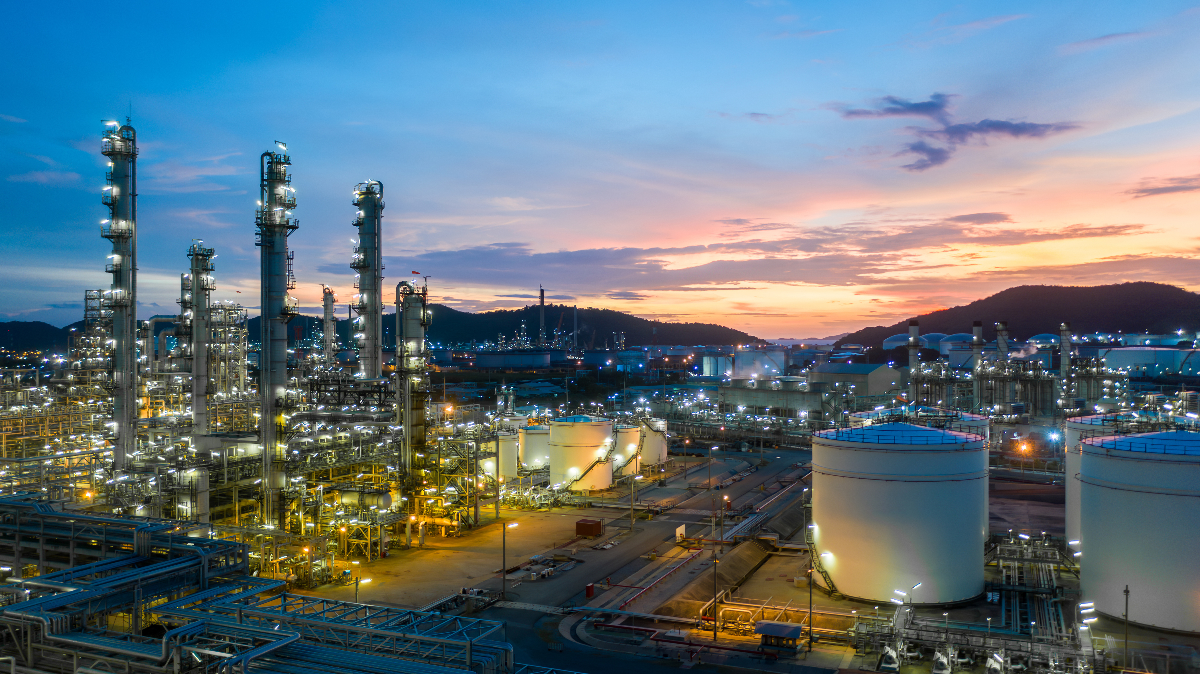 Industrial petrochemical refinery complex illuminated at sunset with tall distillation towers and storage tanks against twilight sky