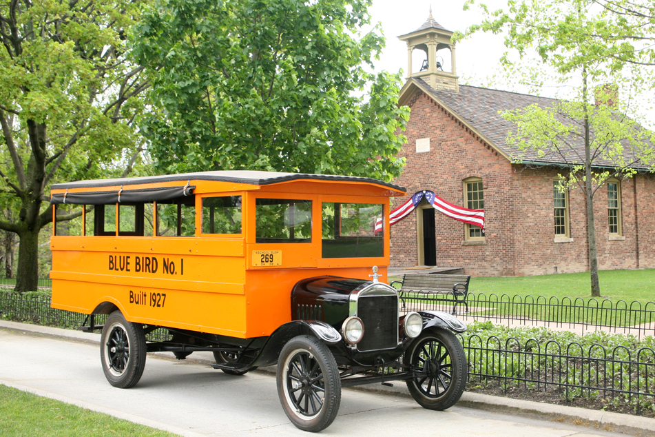 Vintage 1927 Ford Model T truck labeled 'Blue Bird No. 1' parked in front of a brick schoolhouse with bell tower