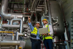 Two industrial workers in safety gear inspecting large HVAC piping systems in a mechanical facility