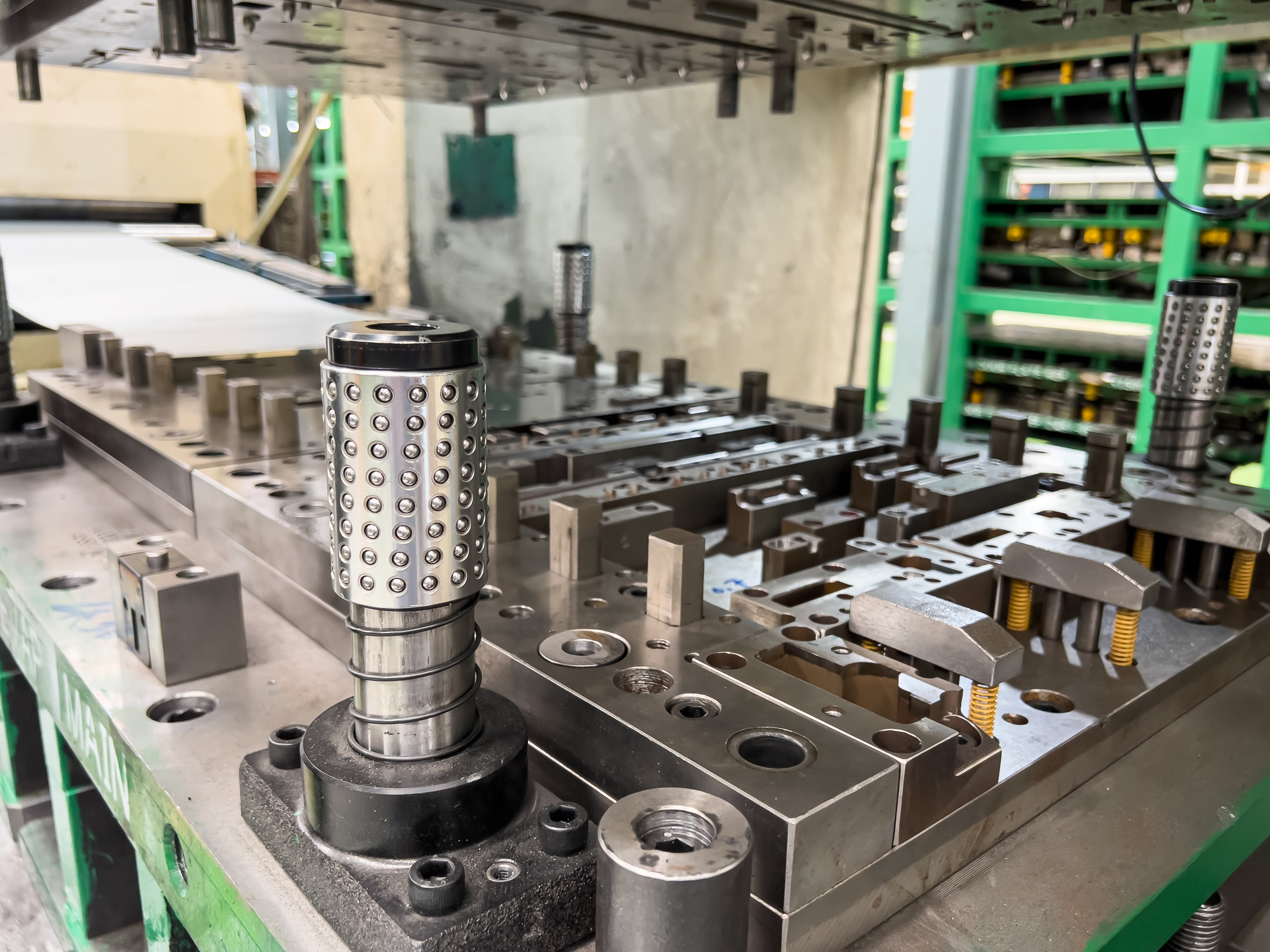 Cylindrical metal part with hole pattern on industrial workbench surrounded by manufacturing equipment and green electronic racks