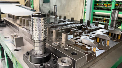 Cylindrical metal part with hole pattern on industrial workbench surrounded by manufacturing equipment and green electronic racks