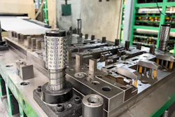 Cylindrical metal part with hole pattern on industrial workbench surrounded by manufacturing equipment and green electronic racks