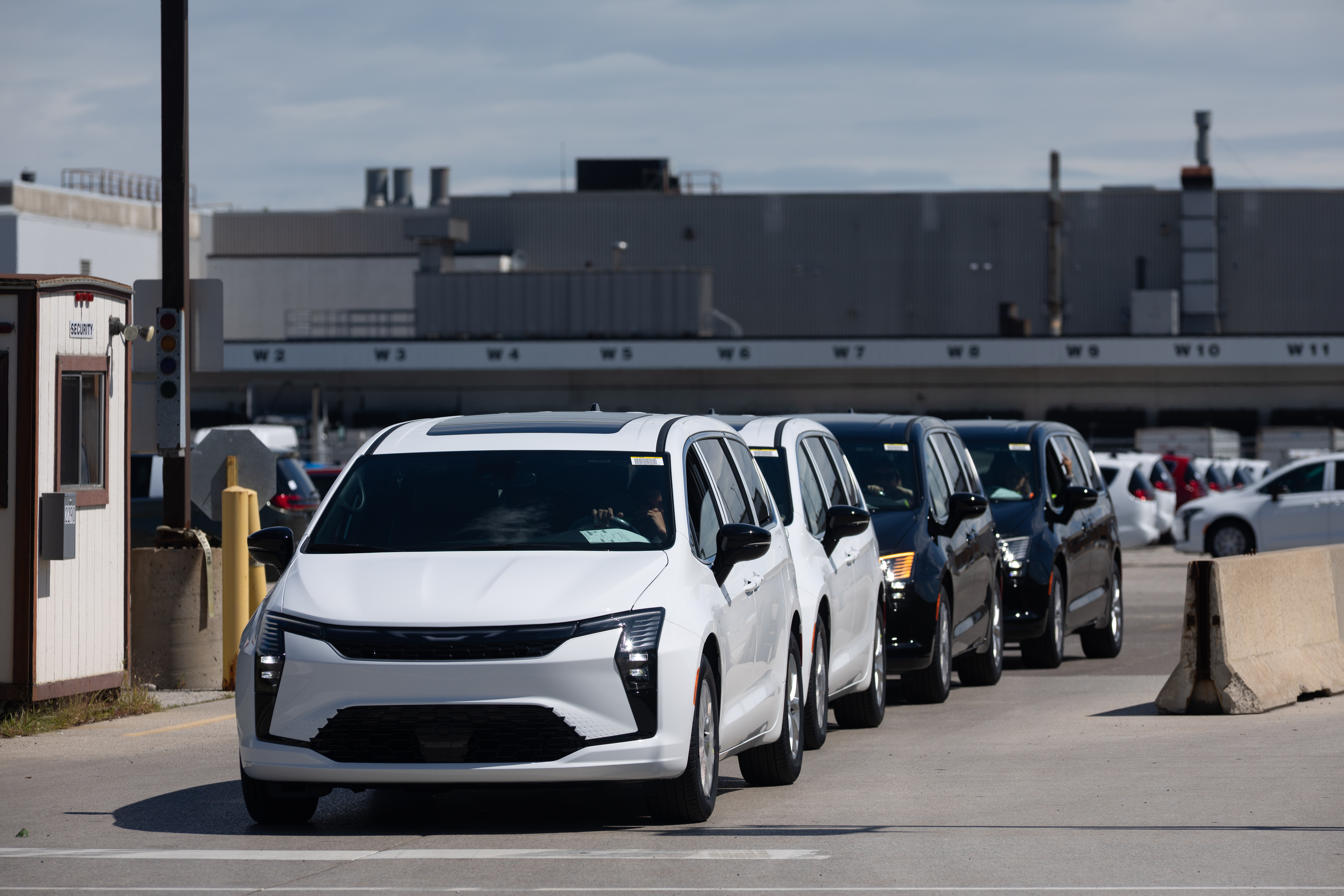 Row of white and dark vehicles parked in industrial lot with factory building in background