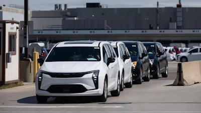 Row of white and dark vehicles parked in industrial lot with factory building in background