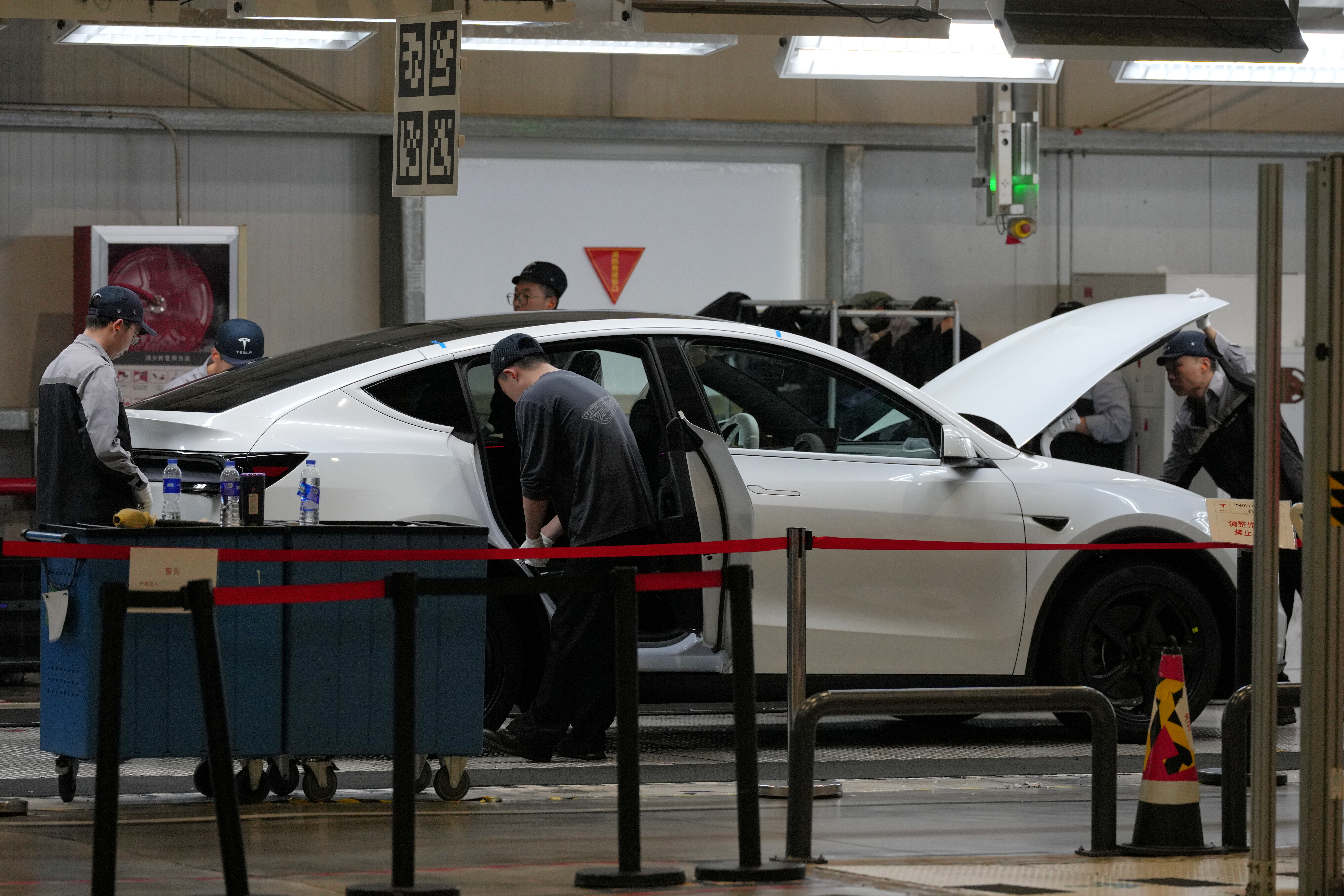 Technicians inspect and service a white sedan in an automotive repair shop with overhead lighting and diagnostic equipment