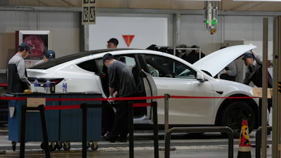 Technicians inspect and service a white sedan in an automotive repair shop with overhead lighting and diagnostic equipment