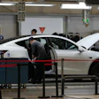 Technicians inspect and service a white sedan in an automotive repair shop with overhead lighting and diagnostic equipment