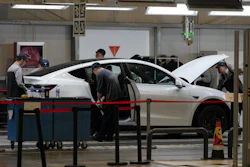 Technicians inspect and service a white sedan in an automotive repair shop with overhead lighting and diagnostic equipment