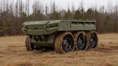 Olive green six-wheeled military armored transport vehicle on muddy terrain with bare trees in background