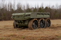 Olive green six-wheeled military armored transport vehicle on muddy terrain with bare trees in background