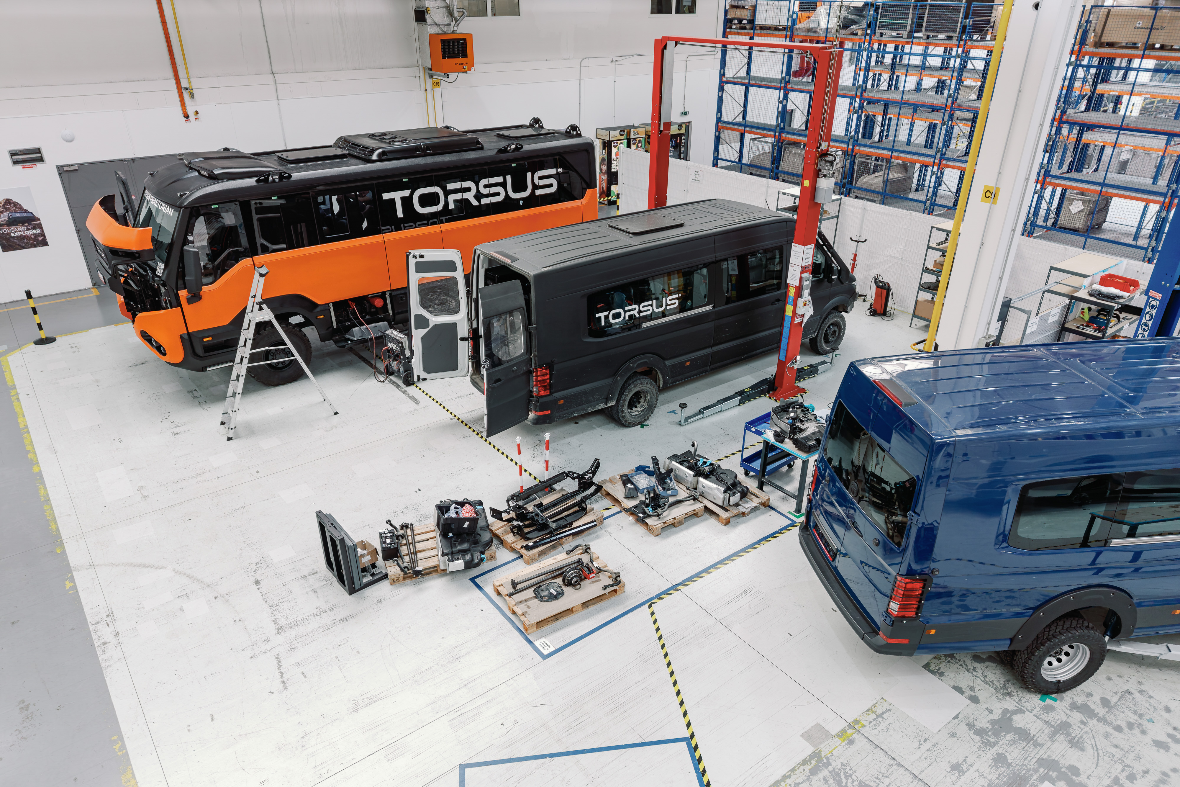 Torsus vehicles and components displayed in industrial workshop facility with orange and black buses, blue van, and mechanical parts.