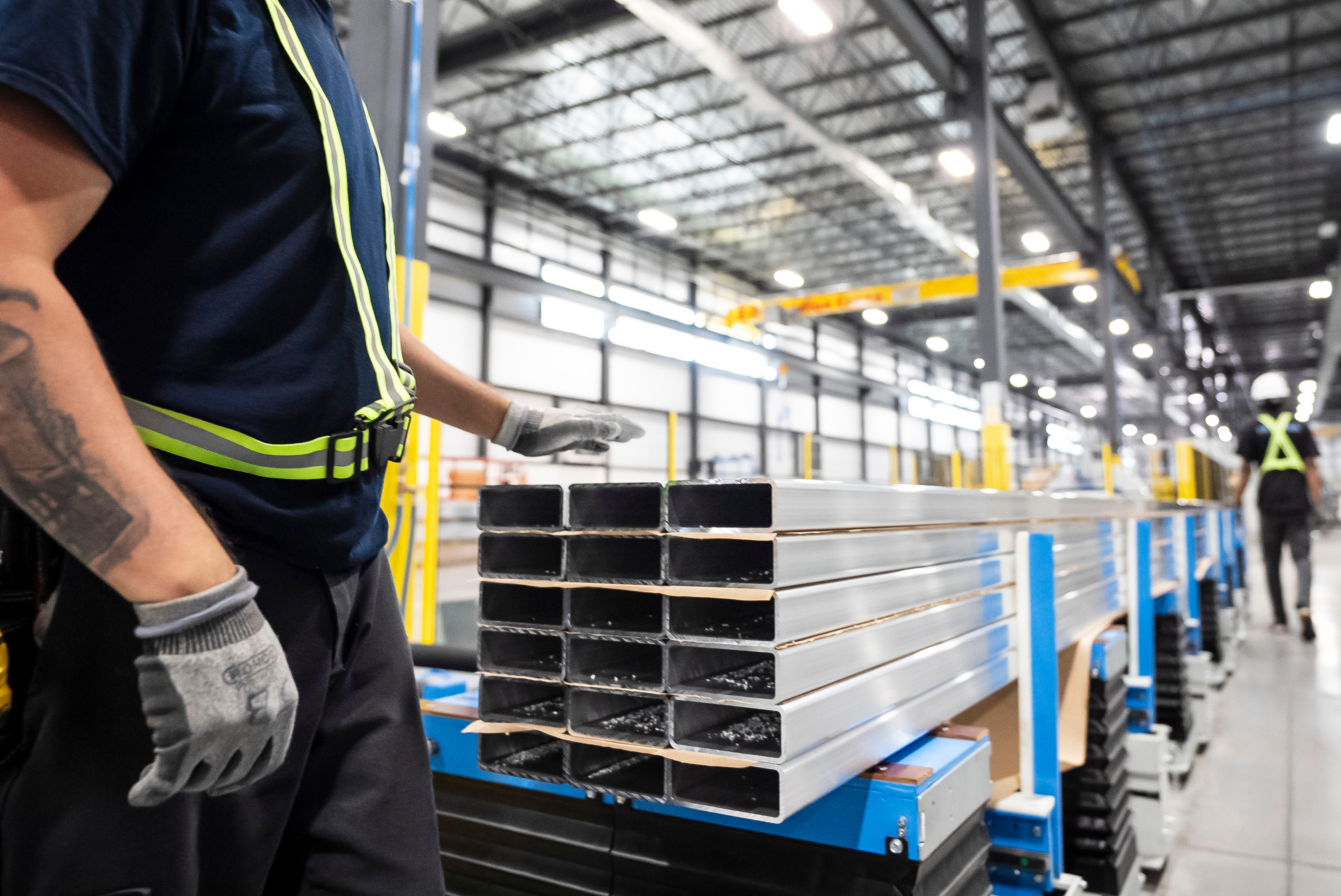 Industrial warehouse worker in safety gear inspecting stacked metal rectangular tubes on blue pallets