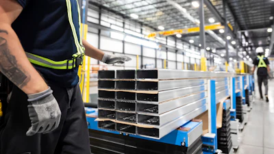 Industrial warehouse worker in safety gear inspecting stacked metal rectangular tubes on blue pallets