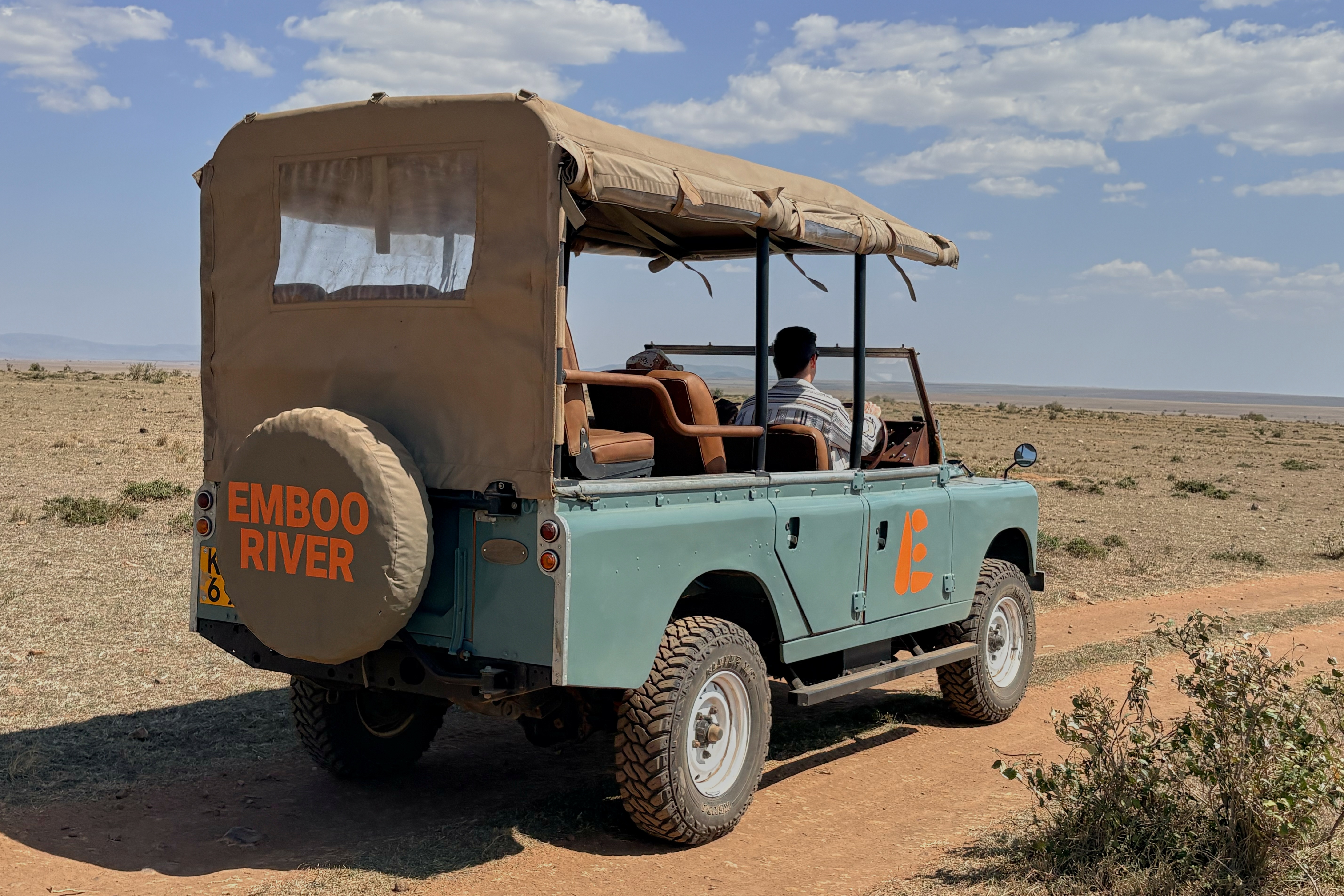 Turquoise safari vehicle with canvas canopy and 'Emboo River' branding parked in African savanna landscape