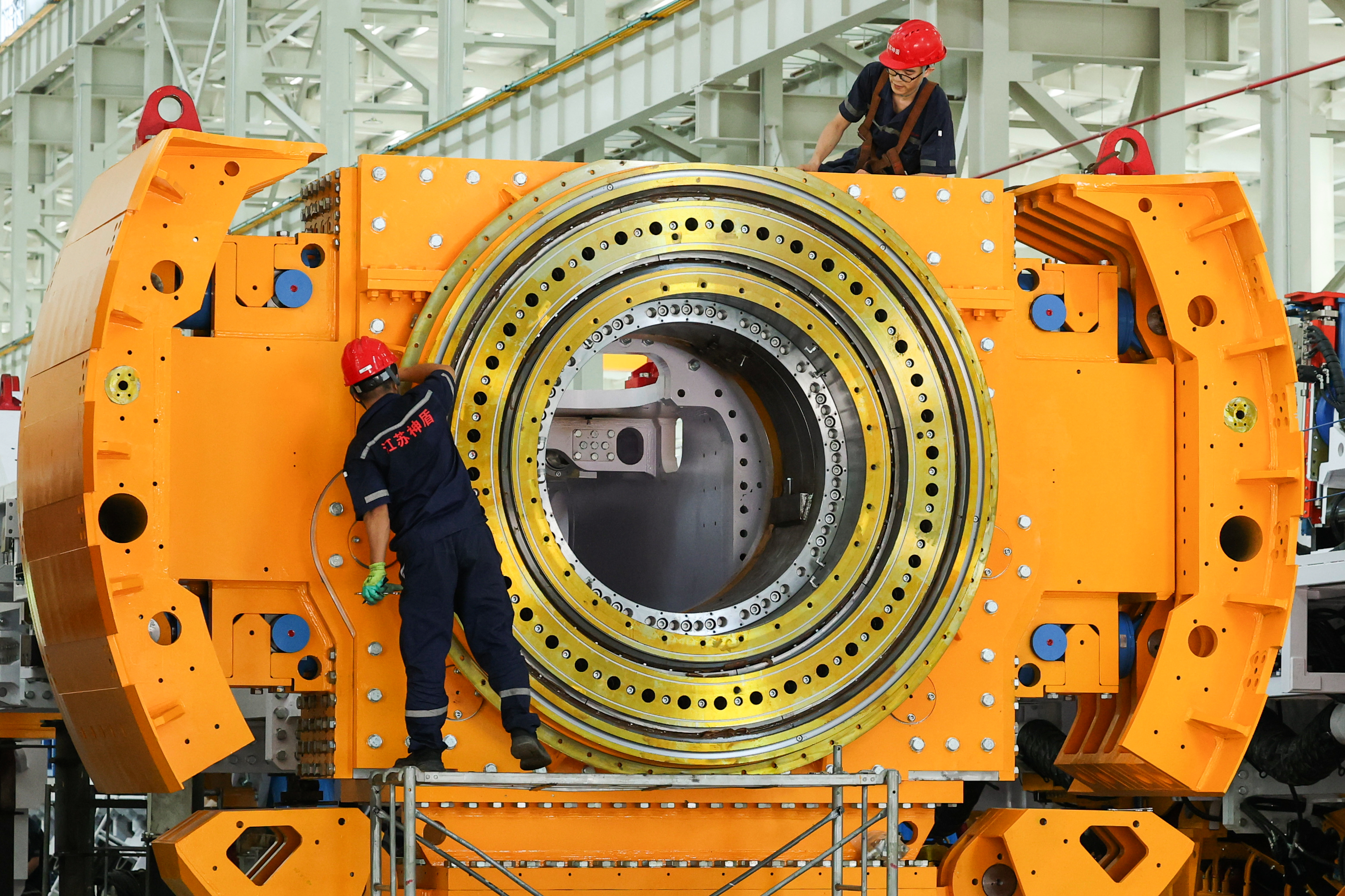 Industrial workers assembling large orange cylindrical machinery with concentric metal rings in factory setting