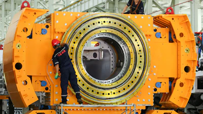 Industrial workers assembling large orange cylindrical machinery with concentric metal rings in factory setting