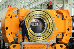 Industrial workers assembling large orange cylindrical machinery with concentric metal rings in factory setting