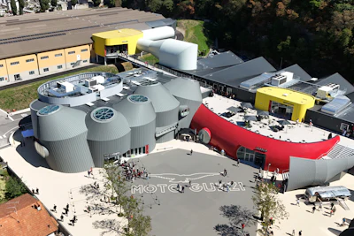 Aerial view of Moto Guzzi industrial facility with cylindrical storage tanks, yellow equipment, and red building structures surrounded by trees.