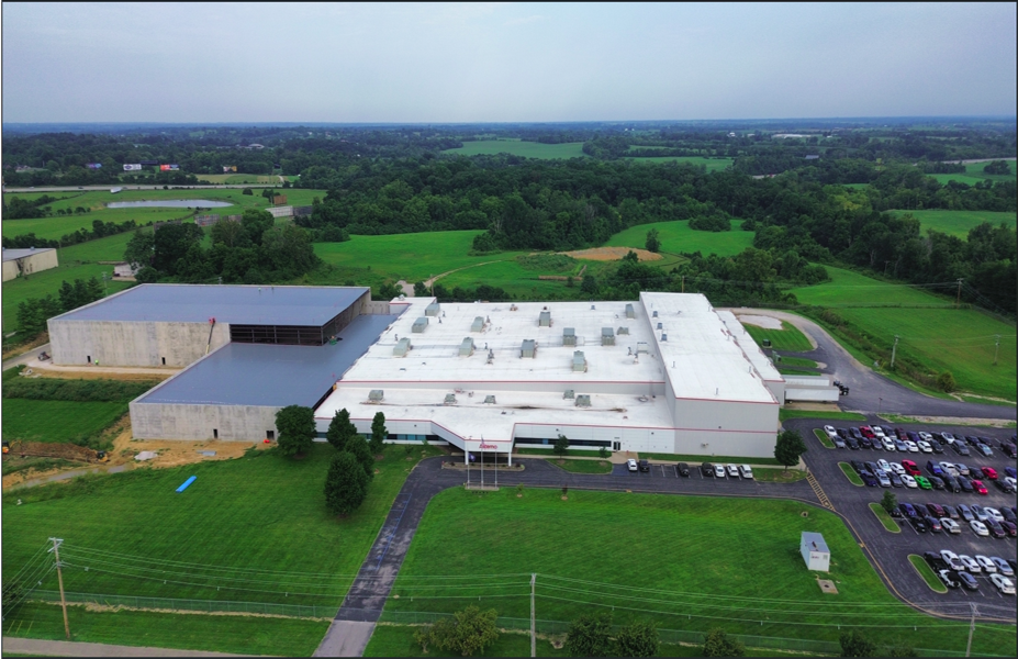 Aerial view of large industrial facility with white-roofed warehouses surrounded by green fields and parking areas