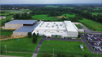 Aerial view of large industrial facility with white-roofed warehouses surrounded by green fields and parking areas