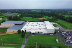 Aerial view of large industrial facility with white-roofed warehouses surrounded by green fields and parking areas