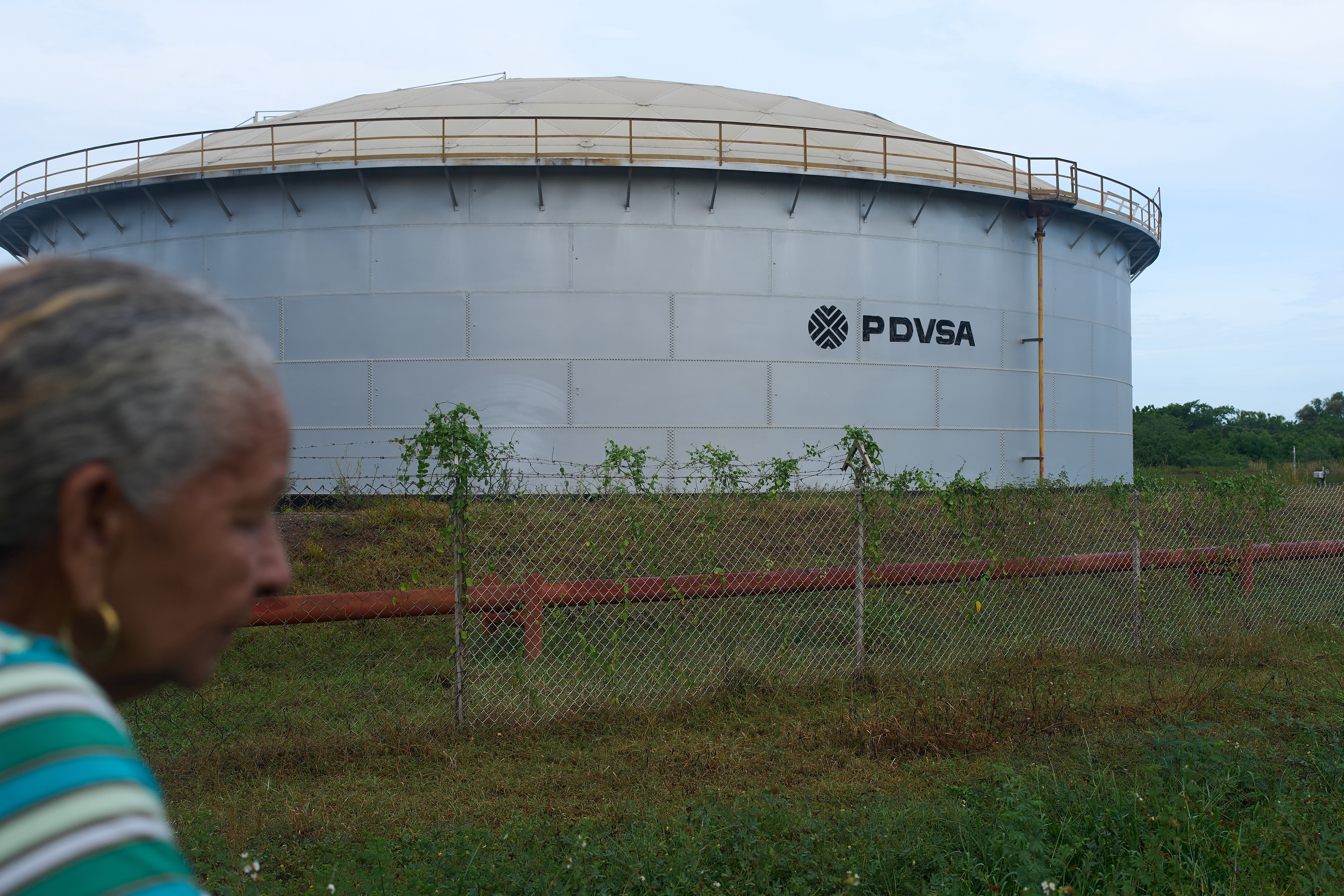 Large cylindrical PDVSA storage tank behind chain-link fence with woman in foreground
