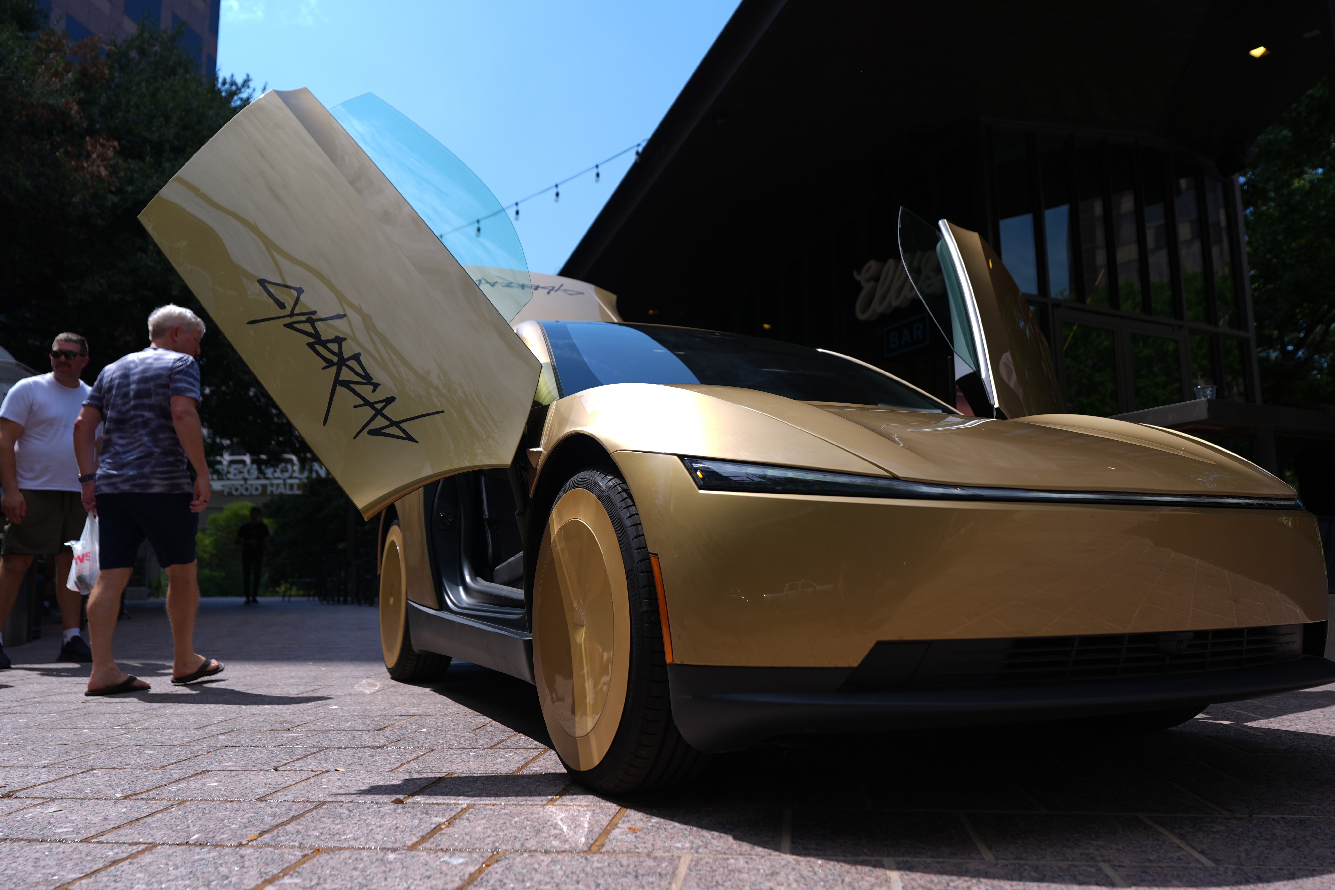 Gold concept car with open gull-wing door displayed at outdoor event with visitors