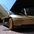 Gold concept car with open gull-wing door displayed at outdoor event with visitors