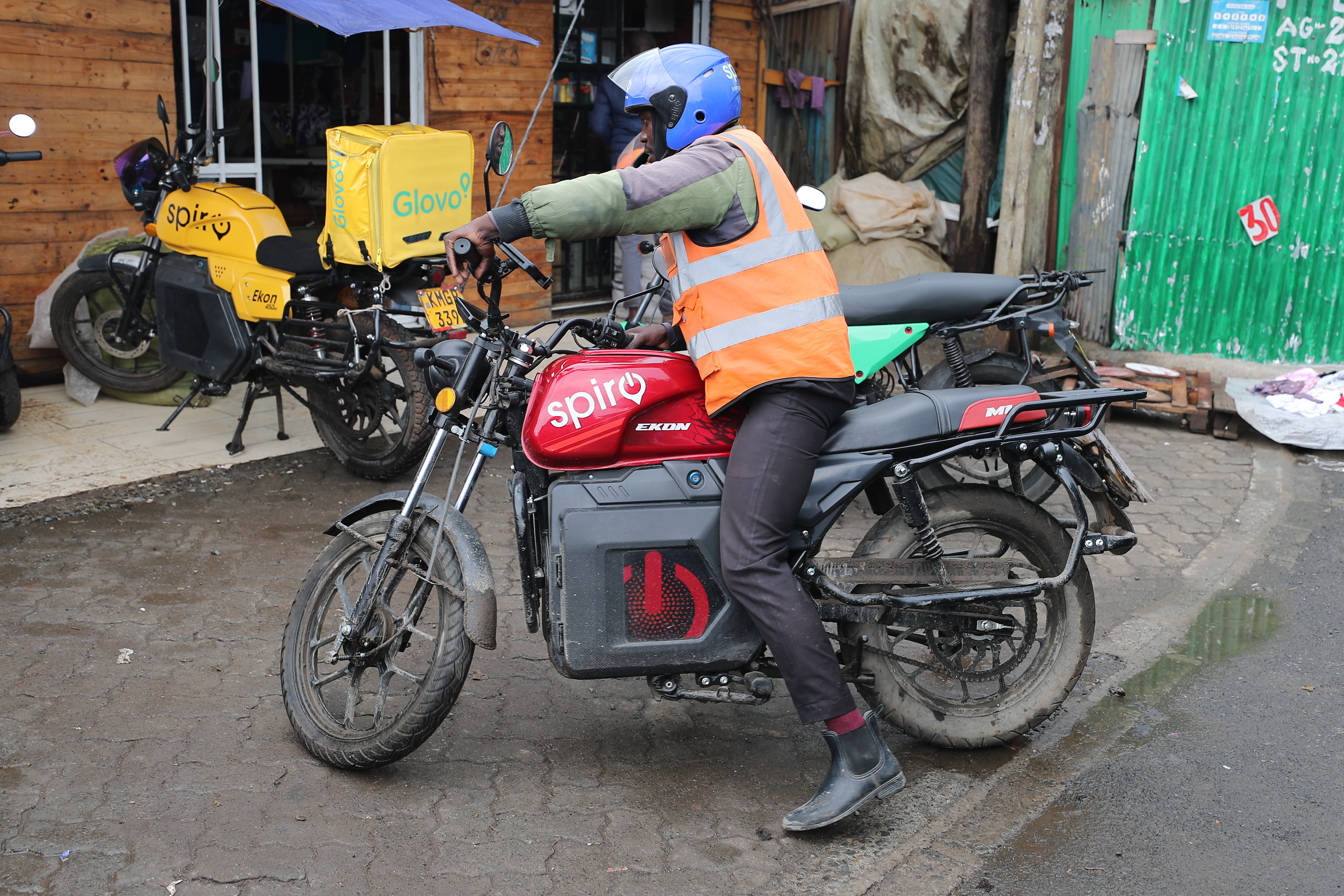 Delivery rider in orange safety vest and blue helmet on Spiro electric motorcycle with delivery box in urban setting