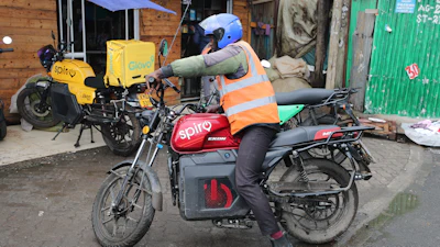 Delivery rider in orange safety vest and blue helmet on Spiro electric motorcycle with delivery box in urban setting