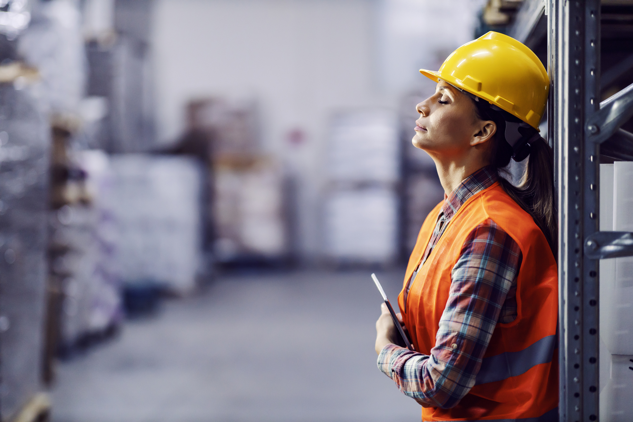 Female warehouse worker in yellow hard hat and orange safety vest holding clipboard in industrial facility