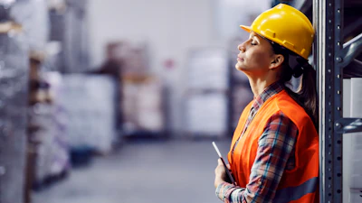 Female warehouse worker in yellow hard hat and orange safety vest holding clipboard in industrial facility