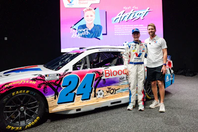 Two men pose beside a colorful NASCAR race car #24 with pink, blue, and wood-grain design at a promotional event