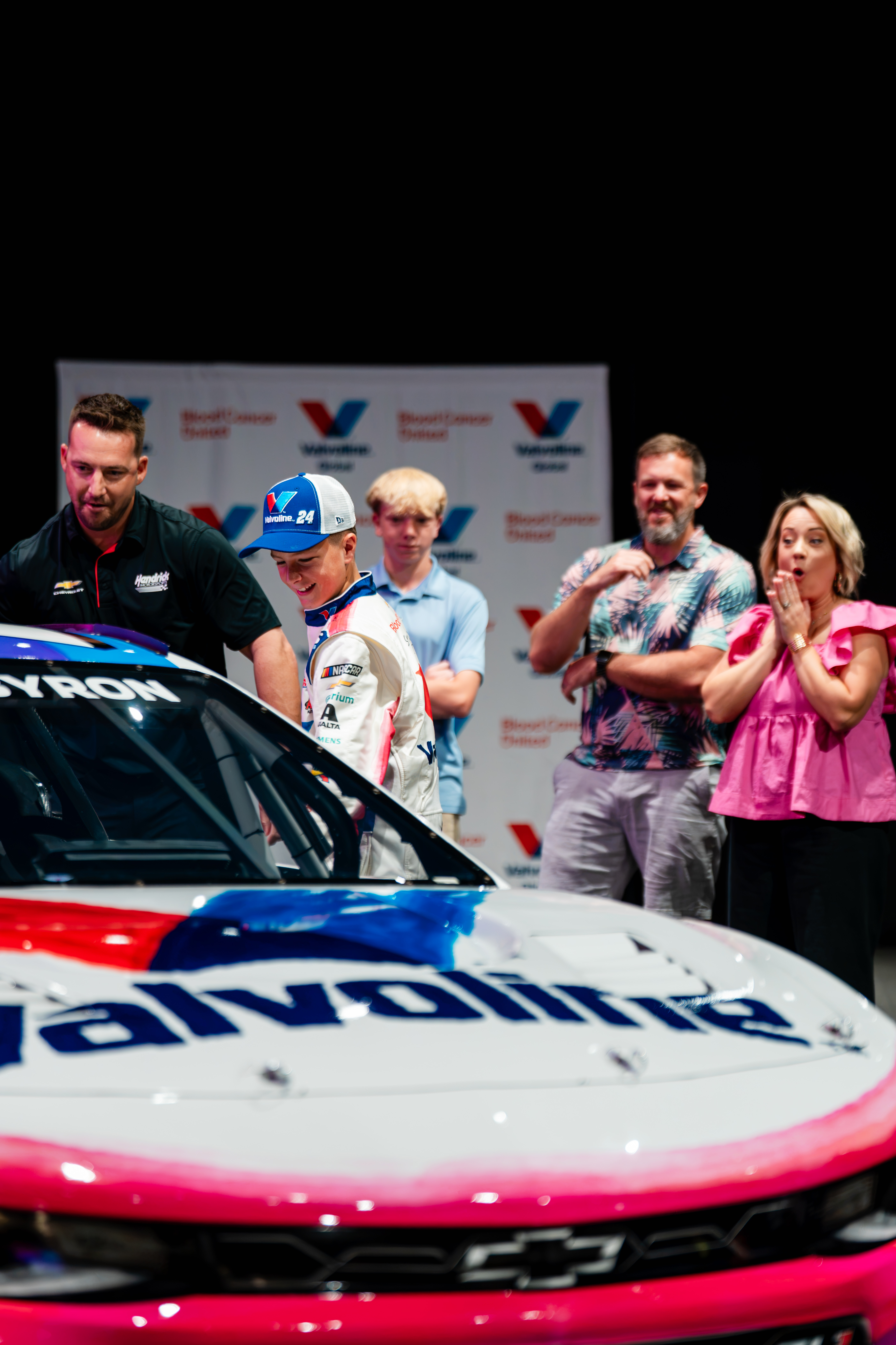 Racing team members and officials gather around a red and white race car during an automotive event presentation.