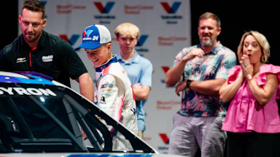 Racing team members and officials gather around a red and white race car during an automotive event presentation.