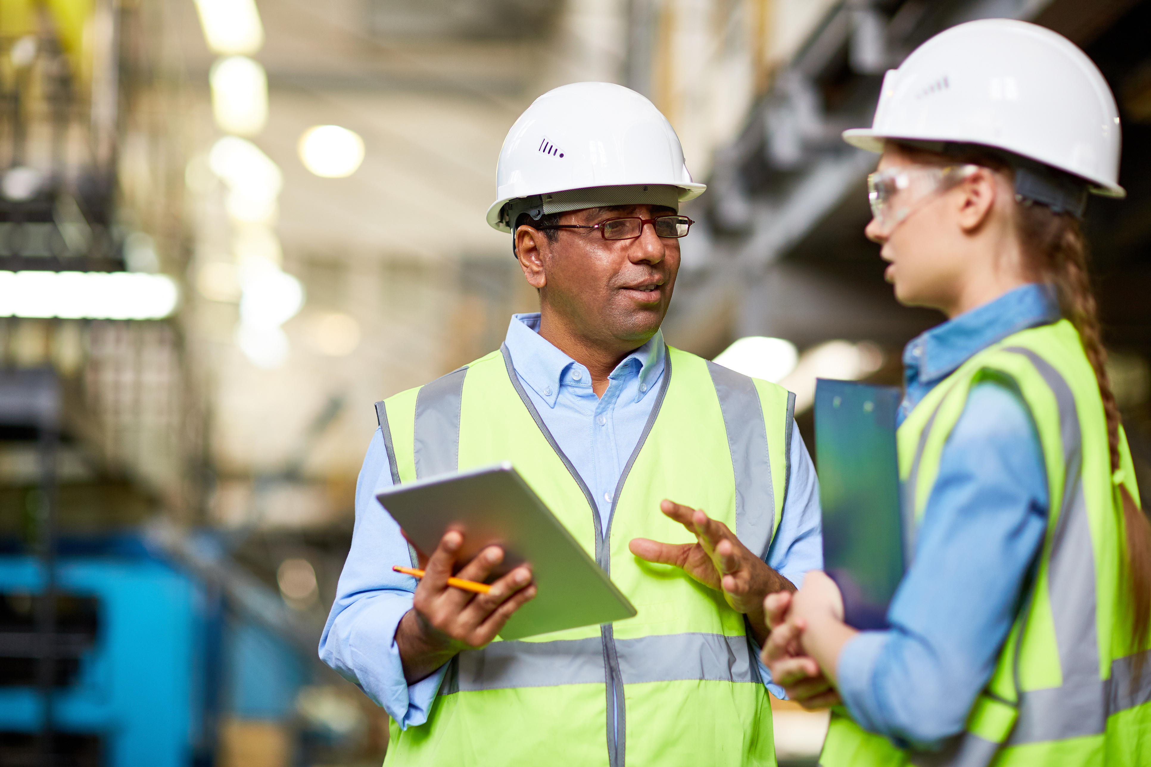 Two industrial workers in hard hats and safety vests review data on a tablet in a factory setting.