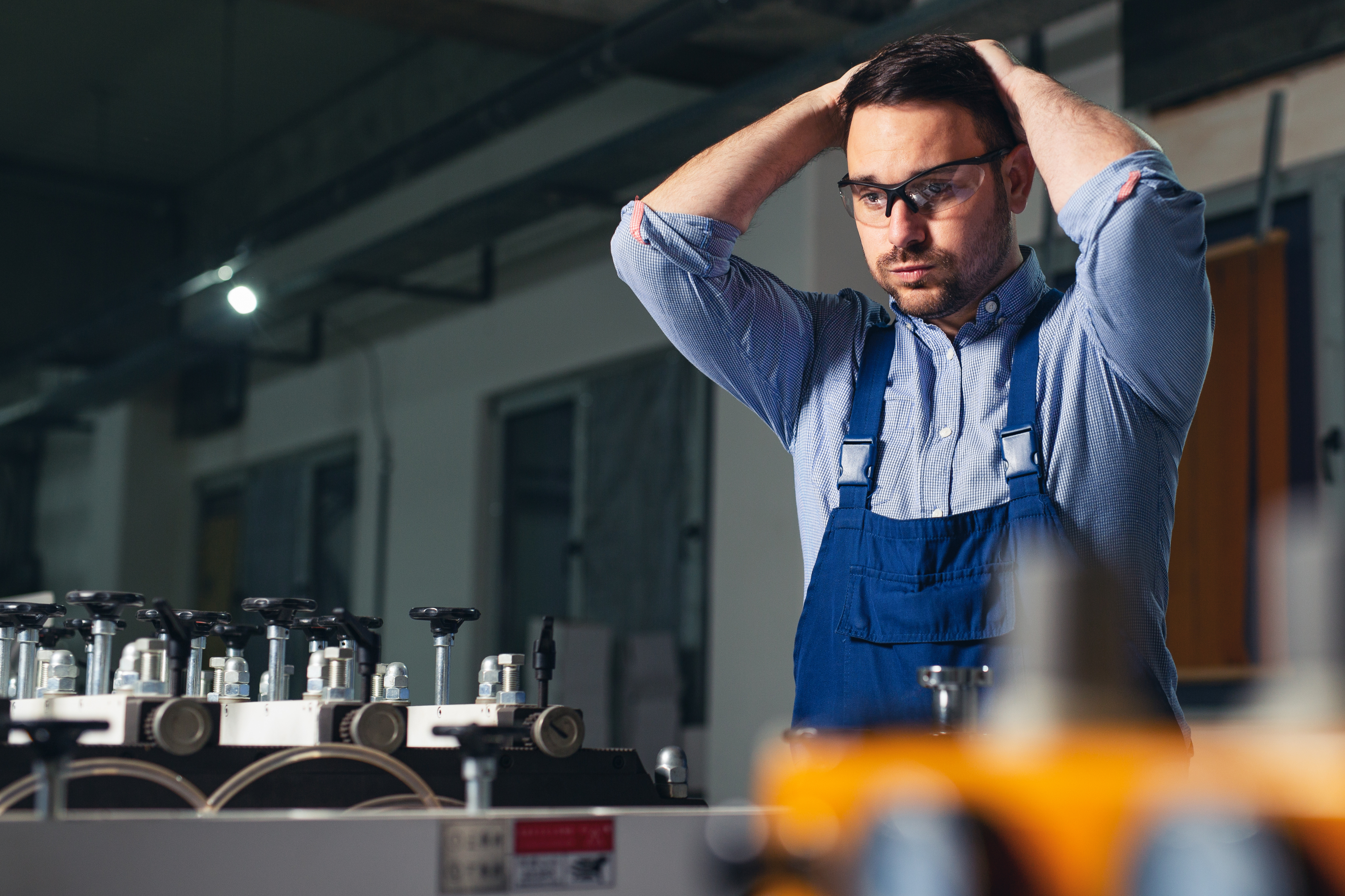 Technician in blue overalls and glasses adjusting his hair in an industrial workshop with equipment on the workbench