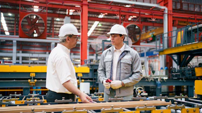 Two workers in hard hats discussing plans at an industrial manufacturing workstation with heavy machinery and equipment