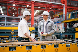 Two workers in hard hats discussing plans at an industrial manufacturing workstation with heavy machinery and equipment