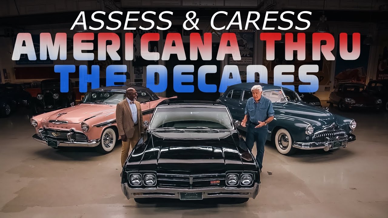 Two men stand with vintage American cars including a pink 1950s convertible and black muscle car in a classic automobile showroom setting