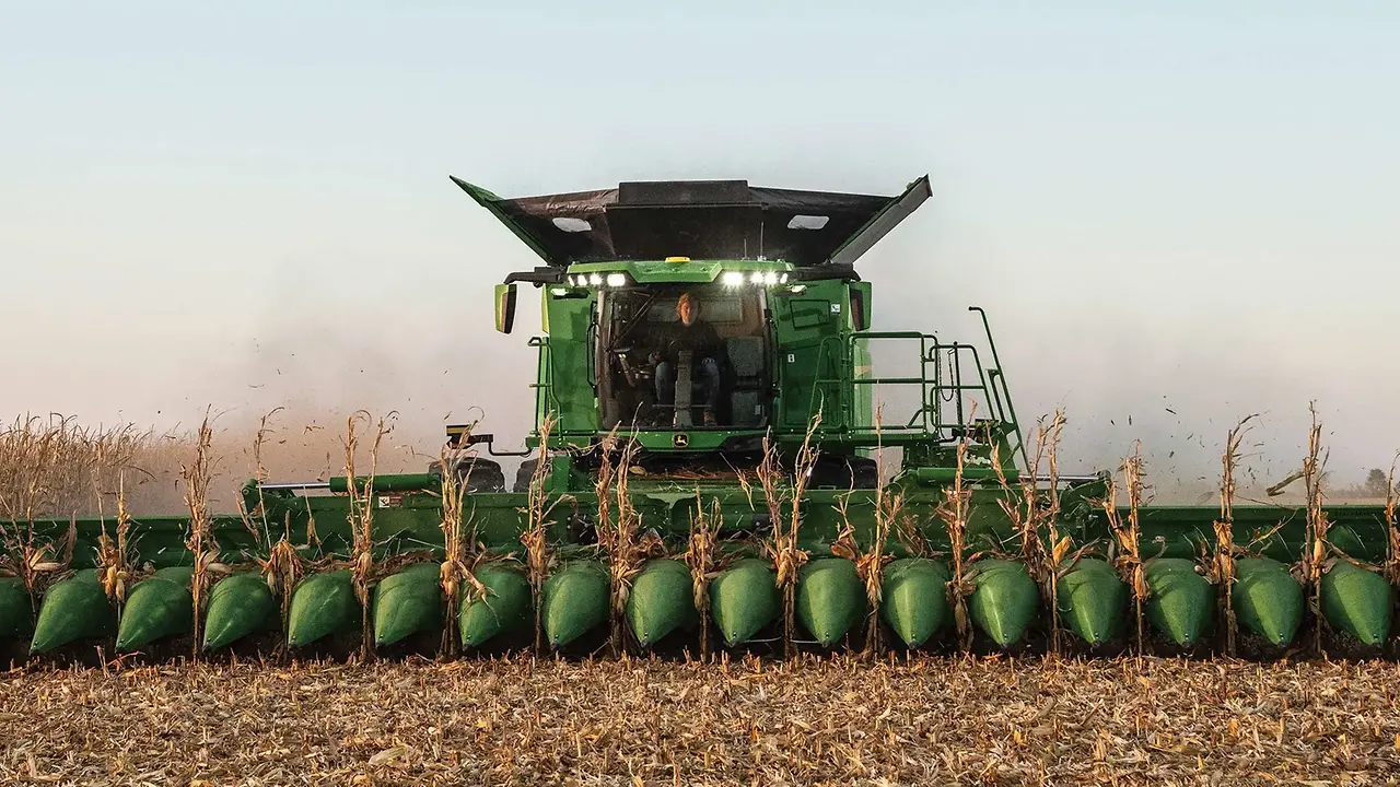 Green John Deere combine harvester with corn header harvesting dried corn stalks in a field at dusk